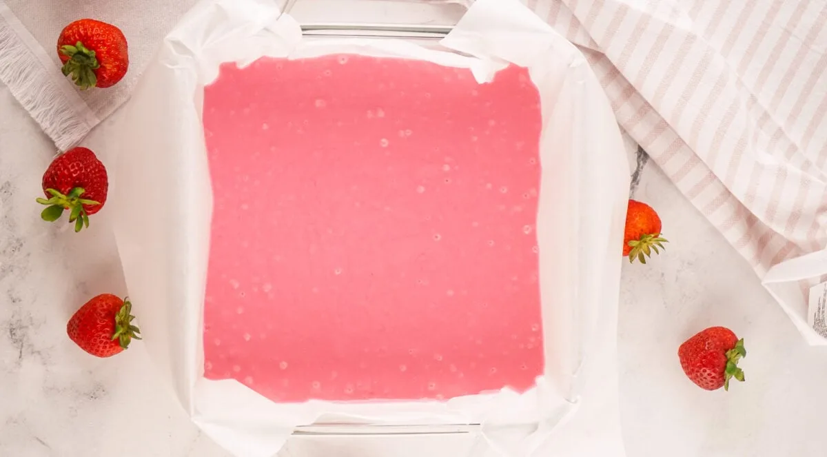 A baking dish lined with parchment paper and filled with pink batter, surrounded by strawberries on a marble countertop.