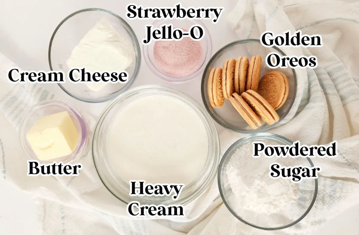 Ingredients for a dessert are displayed: cream cheese, butter, heavy cream, strawberry Jell-O, Golden Oreos, and powdered sugar in separate bowls on a light cloth background.