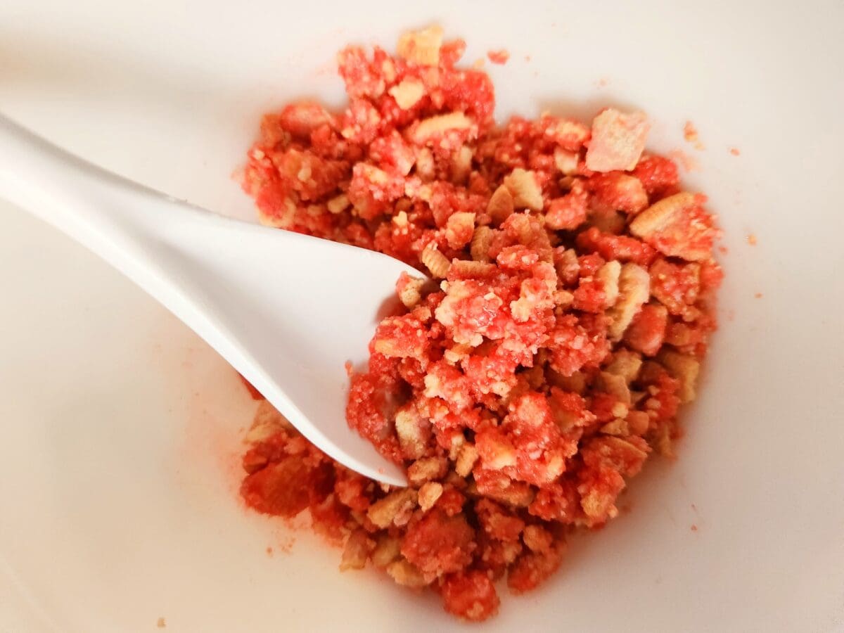 A white bowl with a white spoon holds a mixture of crumbled red and beige crunchy pieces.