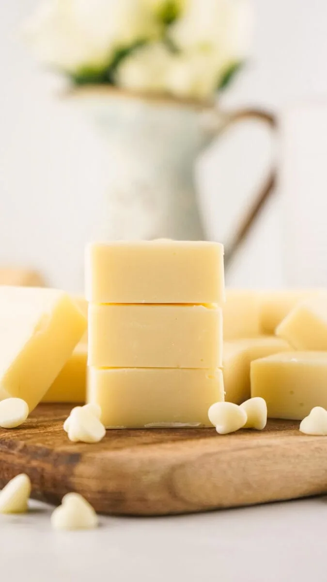 Stacked white chocolate blocks on a wooden board, surrounded by white chocolate chips. Blurred background with a vase of white flowers.