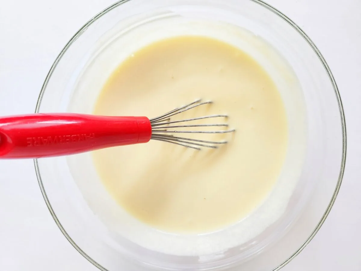 A clear glass bowl filled with creamy batter and a metal whisk with a red handle.