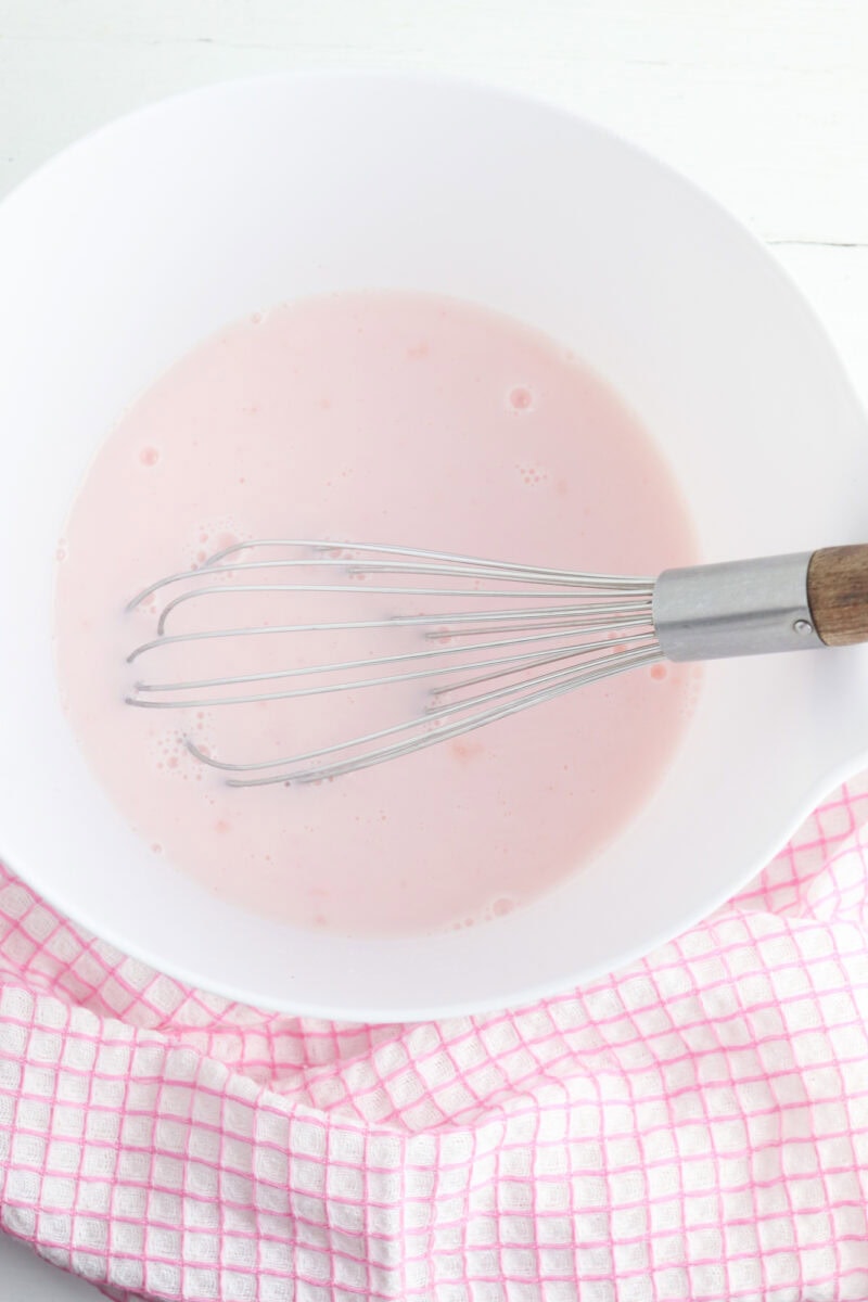 A whisk in a white bowl containing pink liquid, with a pink checkered cloth underneath.