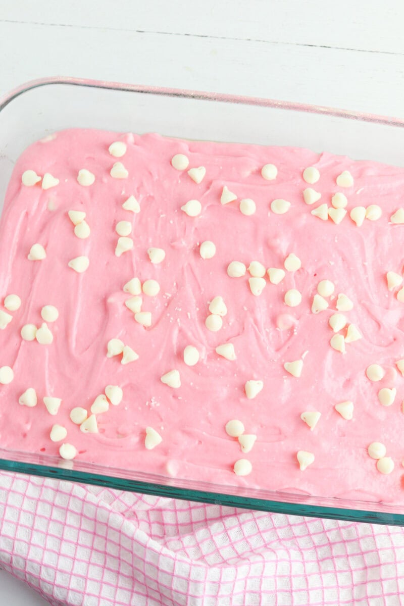 A rectangular glass dish filled with pink frosting topped with white chocolate chips, resting on a pink checkered cloth.