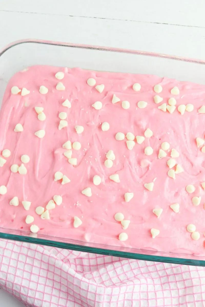 A rectangular glass dish filled with pink frosting topped with white chocolate chips, resting on a pink checkered cloth.