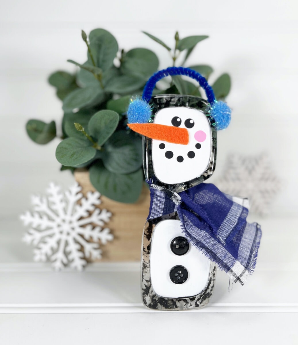 A crafted snowman made of painted blocks, featuring a carrot nose, button eyes, a blue scarf, blue earmuffs, and black buttons, is placed in front of a potted plant and a decorative snowflake.