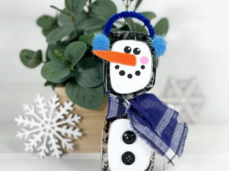 handmade snowman sunglasses craft with a carrot-shaped nose, blue earmuffs, and blue plaid scarf in front of a potted plant and near a decorative white snowflake.