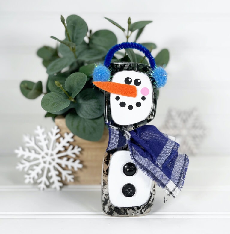 handmade snowman sunglasses craft with a carrot-shaped nose, blue earmuffs, and blue plaid scarf in front of a potted plant and near a decorative white snowflake.