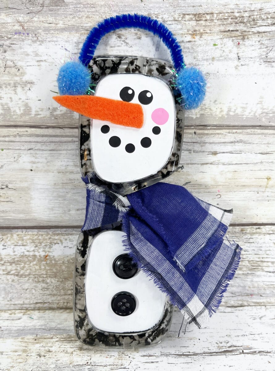 A snowman made from black and white containers, with a felt carrot nose, blue earmuffs, a blue plaid scarf, and two black buttons, is displayed on a wooden surface.