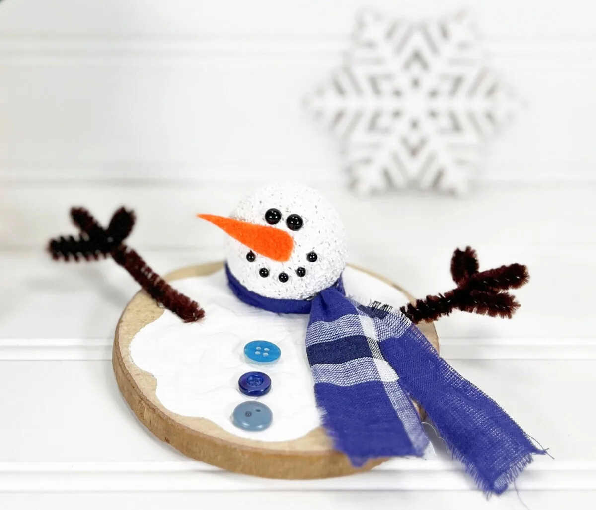 Crafted snowman figure with a blue scarf, orange nose, and button eyes, lying on a wooden base with three blue buttons in front. It has stick-like arms and a blurred snowflake backdrop.