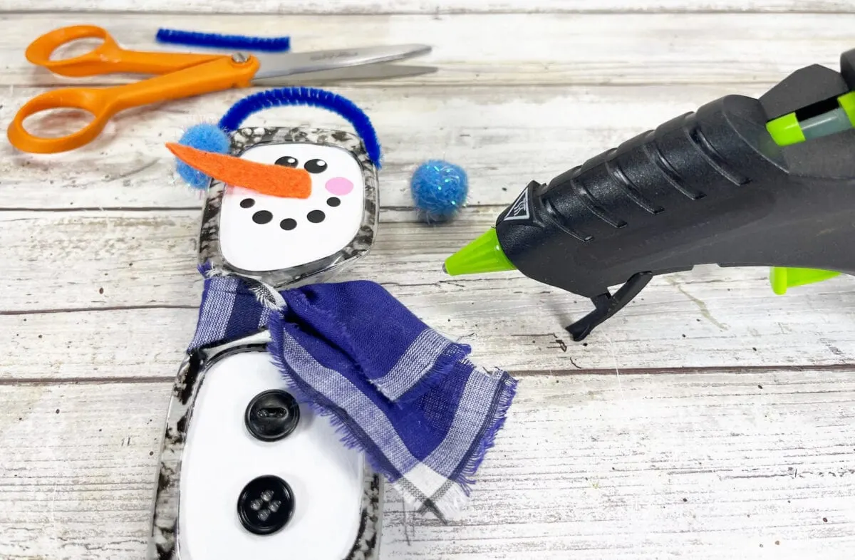 Craft materials and tools on a table: a snowman decoration with a blue scarf, buttons, and an orange felt nose, surrounded by scissors, blue pompoms, and a glue gun.