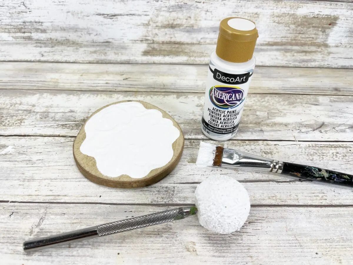 Crafting tools on a wooden surface: a bottle of white acrylic paint, a wooden circle partially painted white, a paintbrush, a small knife, and a textured foam ball.