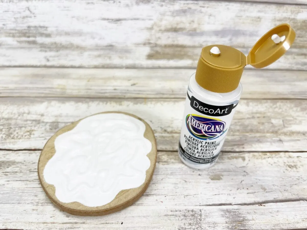 Open bottle of DecoArt Americana acrylic paint next to a wooden slice partially covered in white paint.