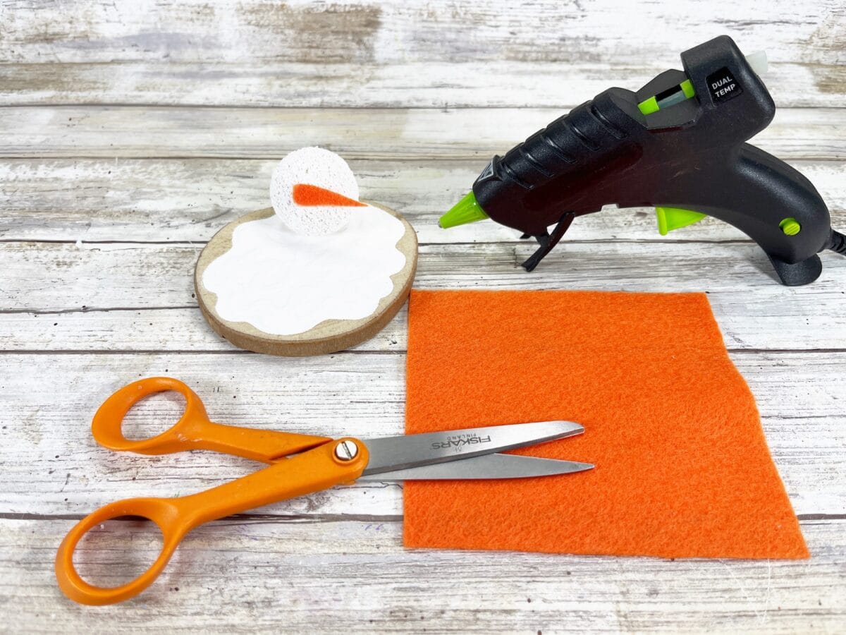 Crafting materials arranged on a white wooden surface, including a hot glue gun, scissors, orange fabric, a round sponge, and a cardboard piece with white glue.