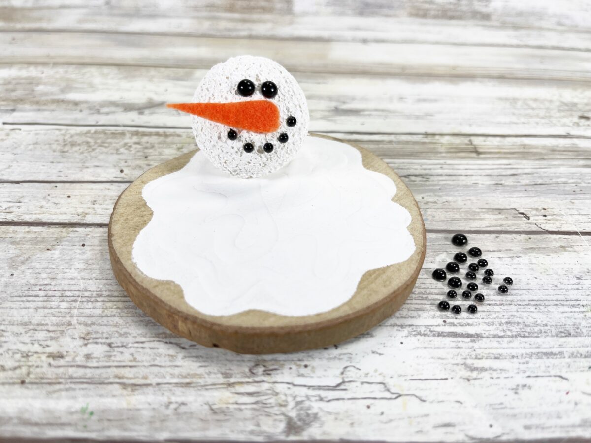 Crocheted snowman head with orange nose on a wooden base, surrounded by small black beads, on a wooden surface.