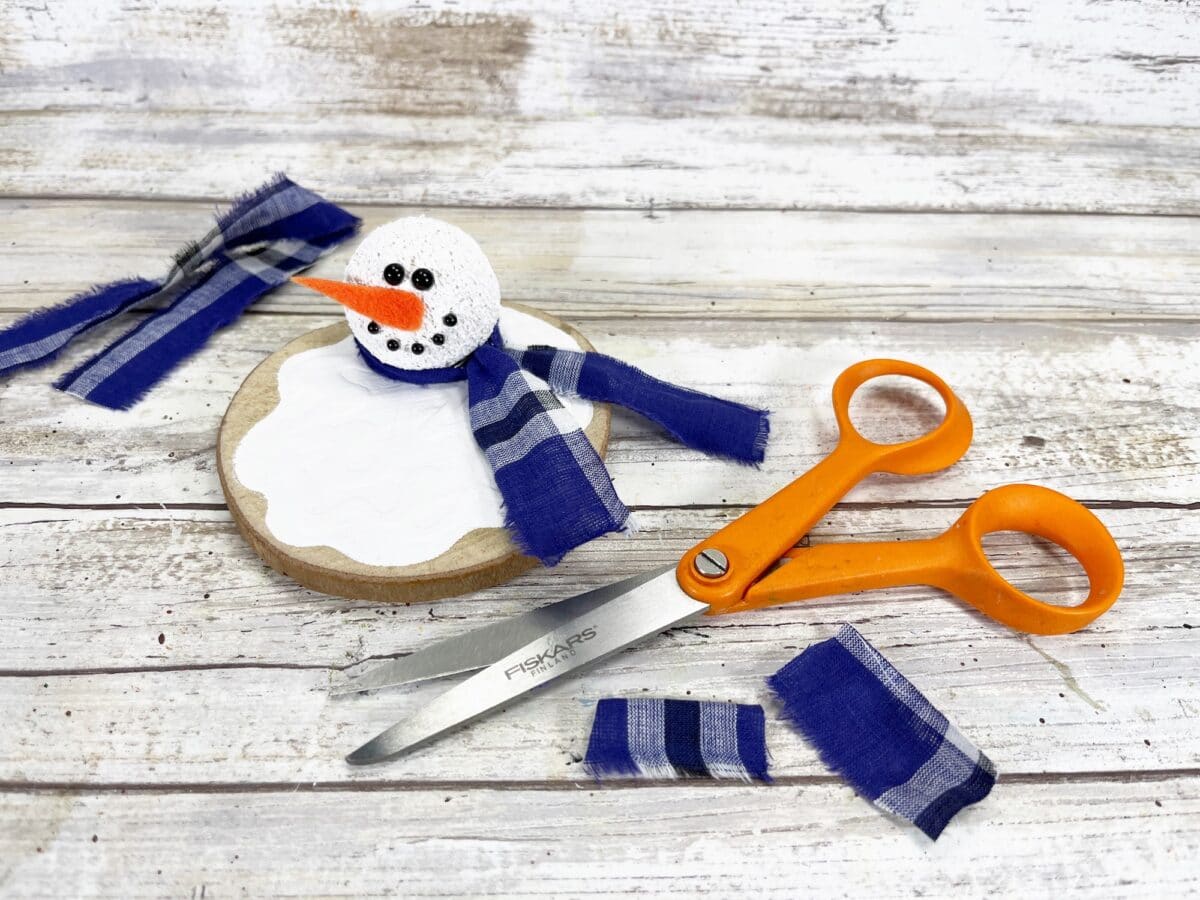 Crafting scene with a small snowman on a wooden base, wearing a blue checkered scarf. Orange scissors and fabric pieces are on a white wooden background.