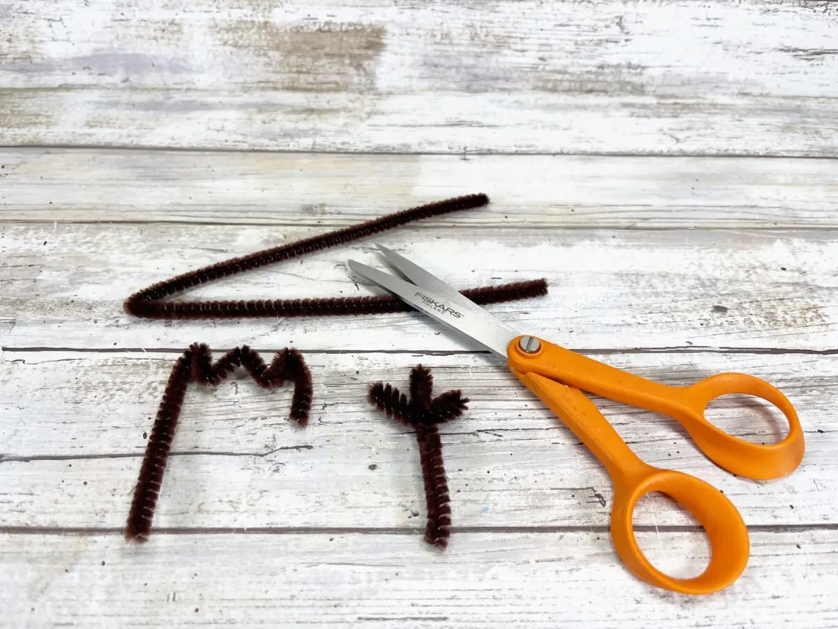 Orange scissors next to cut brown pipe cleaners forming a triangle, letter "M," and an arrow on a wooden surface.