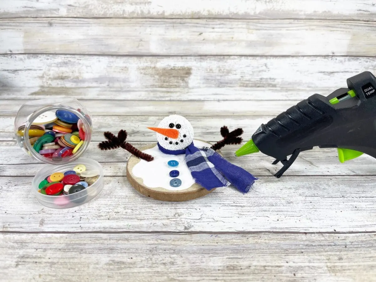 Snowman craft with a fabric scarf and button decorations on a wooden base, next to a glue gun and a container of assorted buttons.