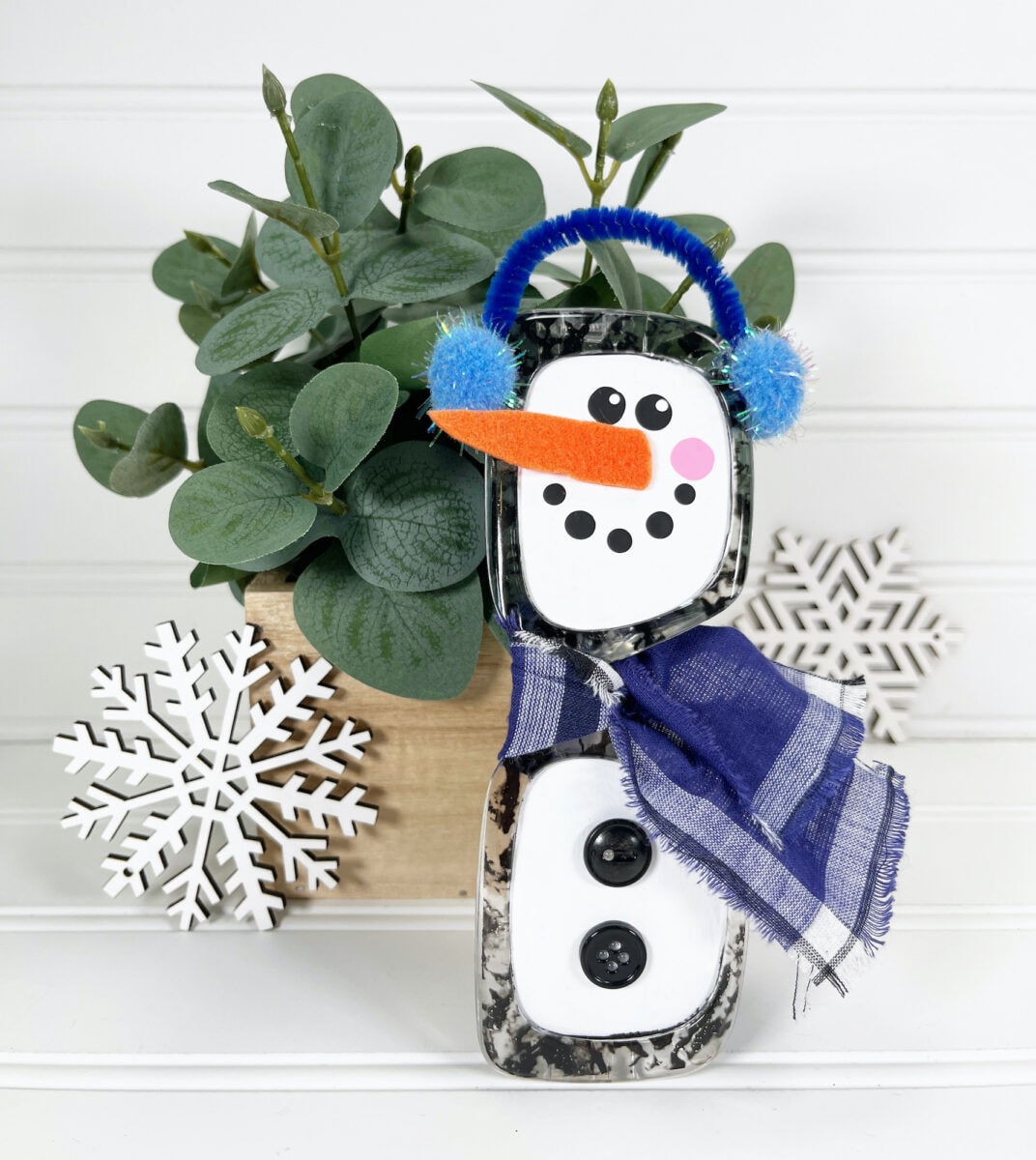 A ceramic snowman with blue earmuffs and a scarf stands beside a potted plant and two decorative white snowflakes on a white background.