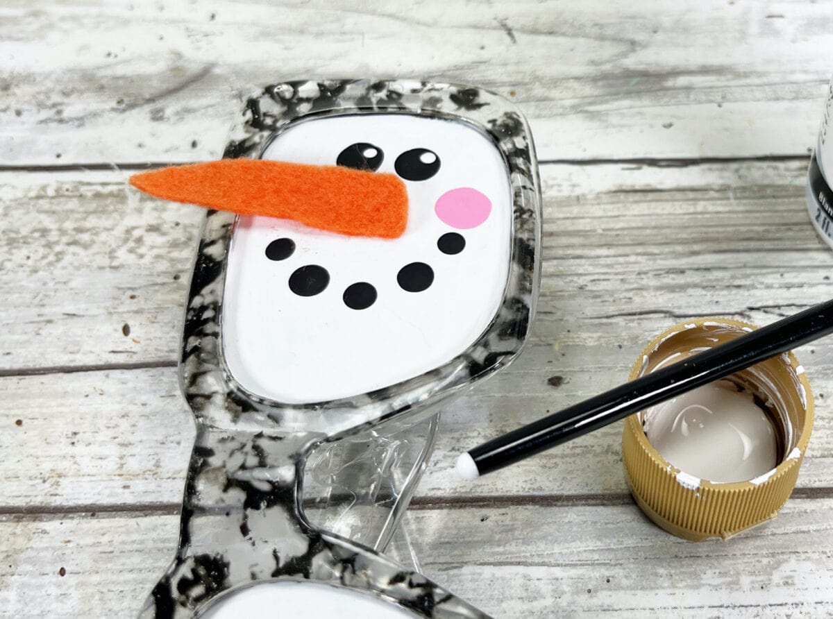 Sunglasses with a snowman face design, including orange felt nose, black eyes and mouth, on a wooden surface with a black pen and paint lid nearby.