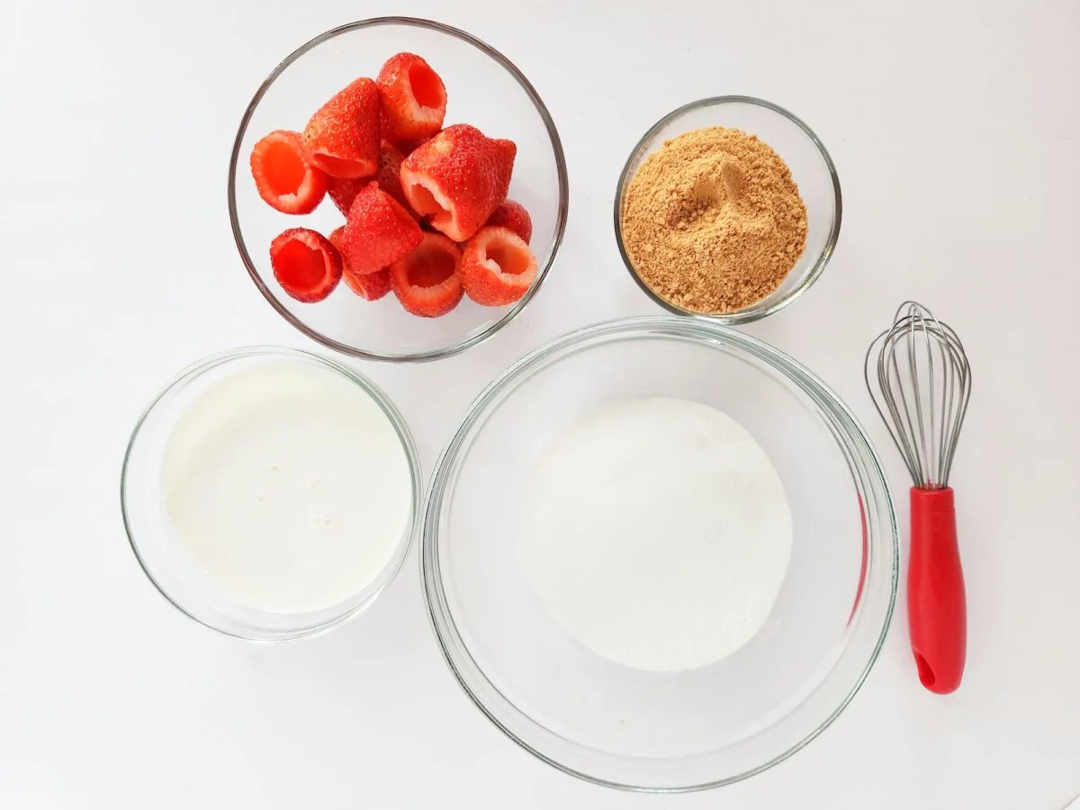 Glass bowls containing cheesecake stuffed strawberries, brown sugar, milk, and white sugar are arranged on a white surface next to a whisk with a red handle.