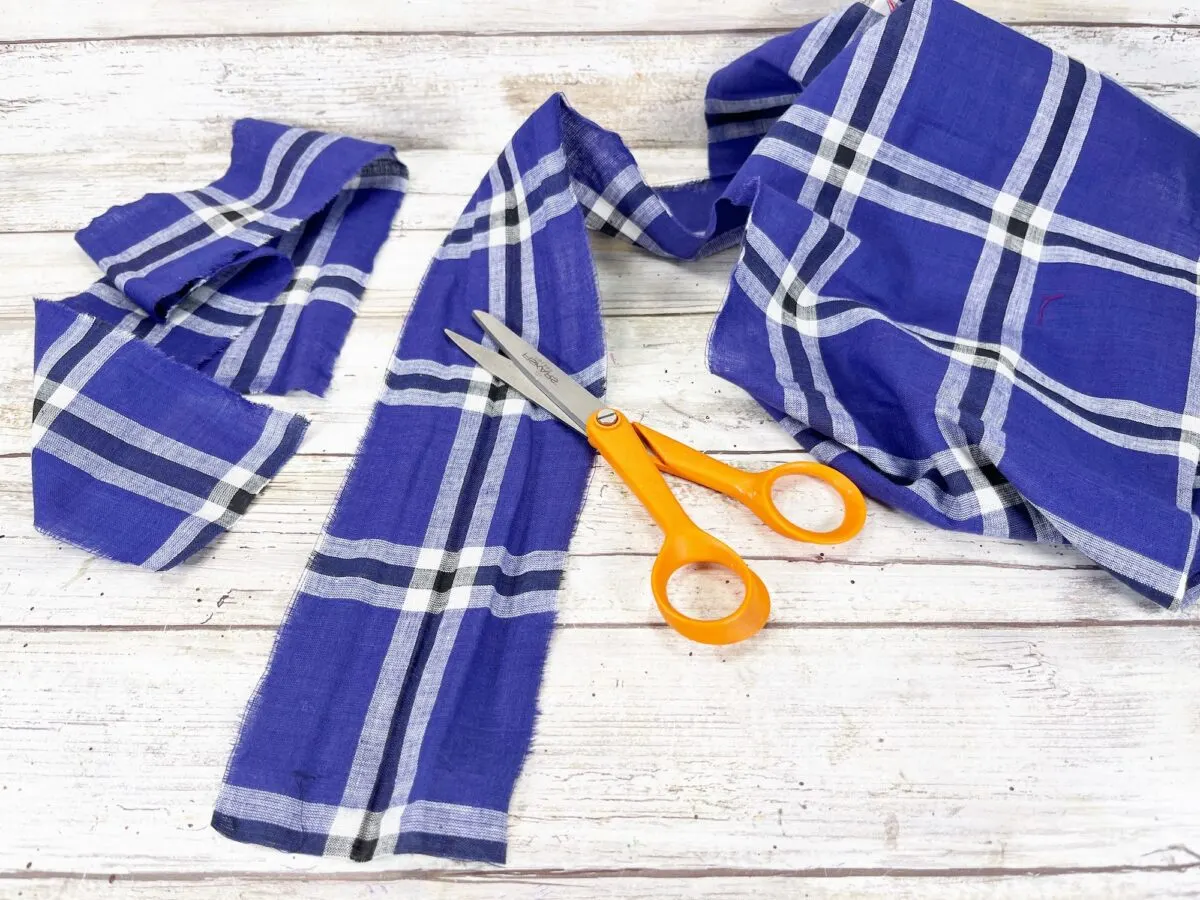 Orange scissors on a wooden surface next to blue plaid fabric pieces, some cut into strips.