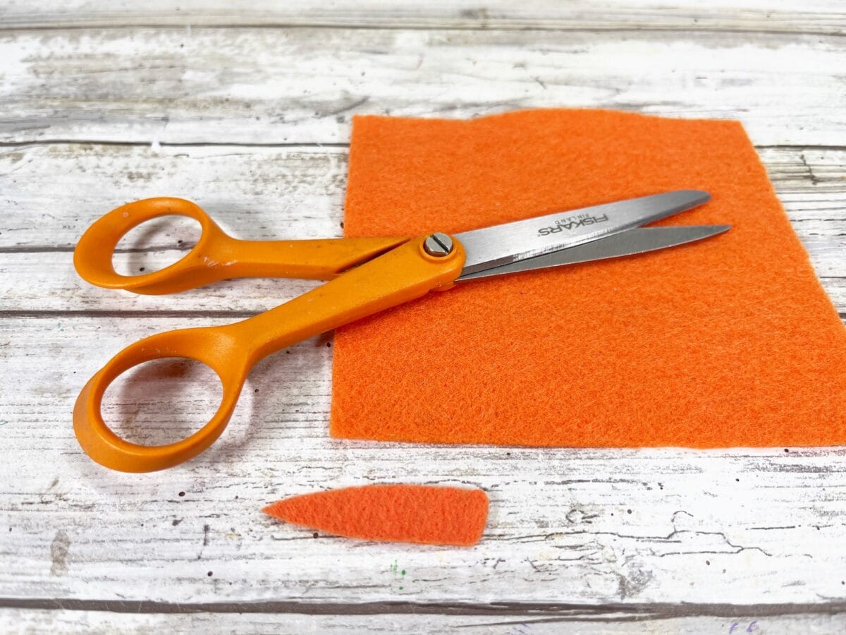 Orange-handled scissors on a wooden surface with an orange felt square and a cut-out triangle piece.