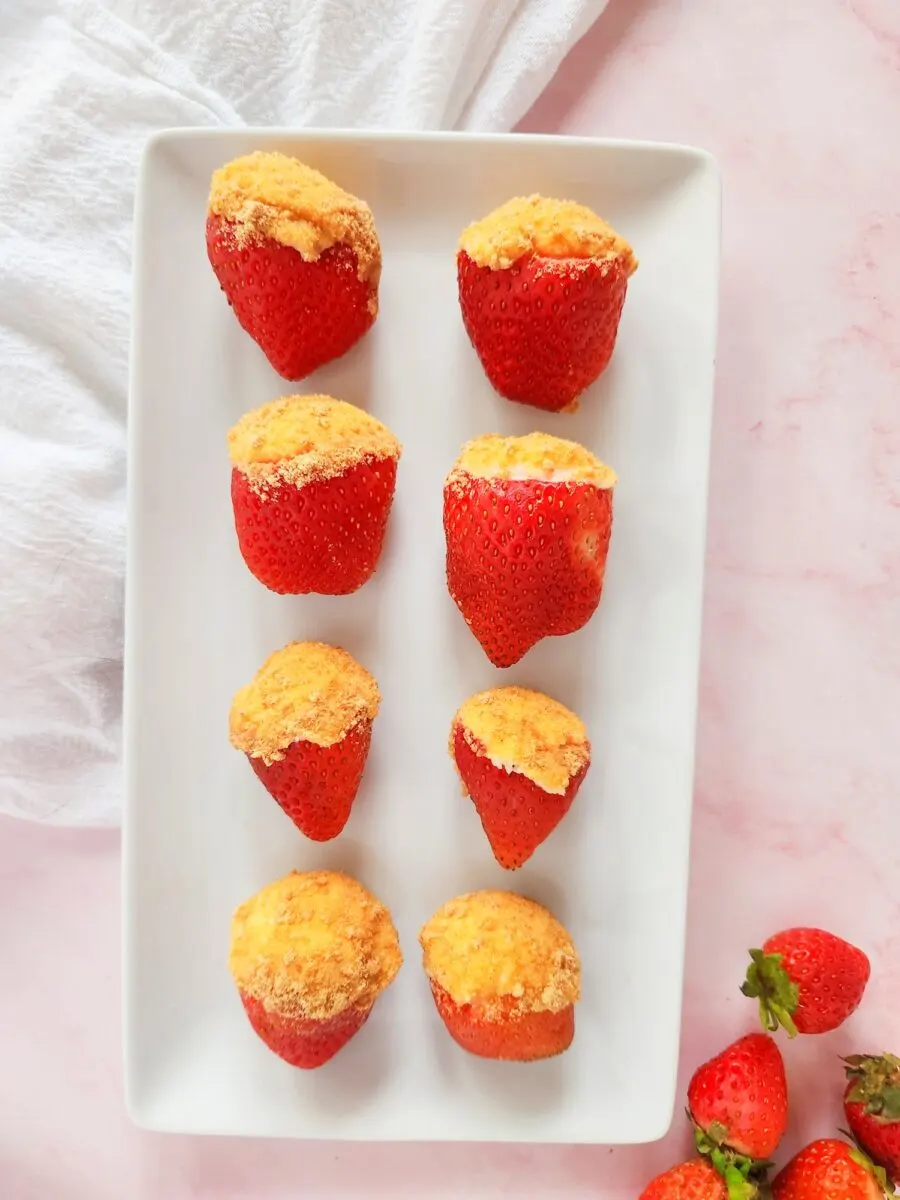 Nine delicious cheesecake-stuffed strawberries are artfully arranged on a rectangular white plate, with a few whole strawberries elegantly placed beside them on a vibrant pink surface.