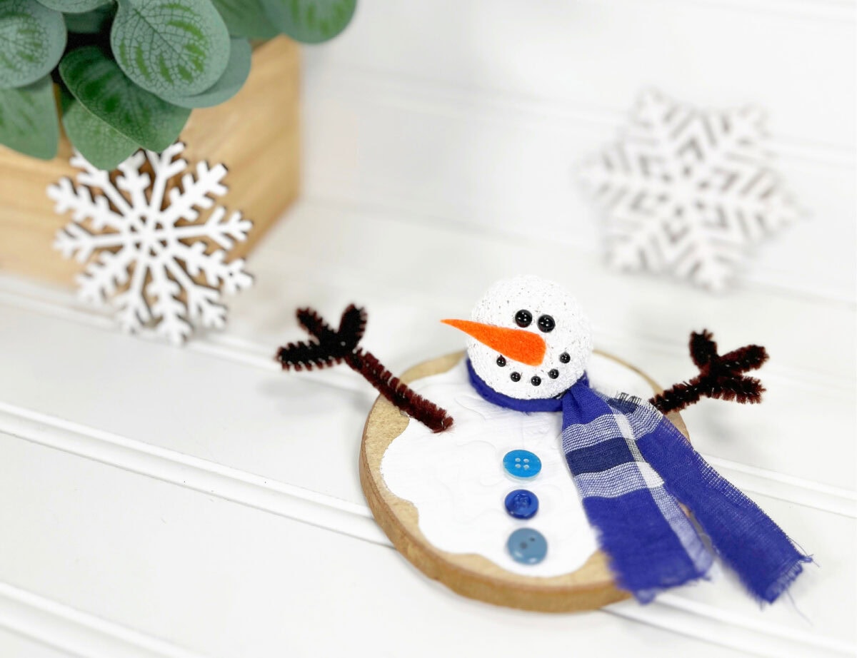 Crafted snowman ornament with a blue scarf, orange carrot nose, and twig-like arms on a wooden base, set against snowflake decorations and a plant.