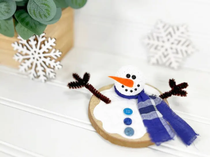 Crafted snowman ornament with a blue scarf, orange carrot nose, and twig-like arms on a wooden base, set against snowflake decorations and a plant.