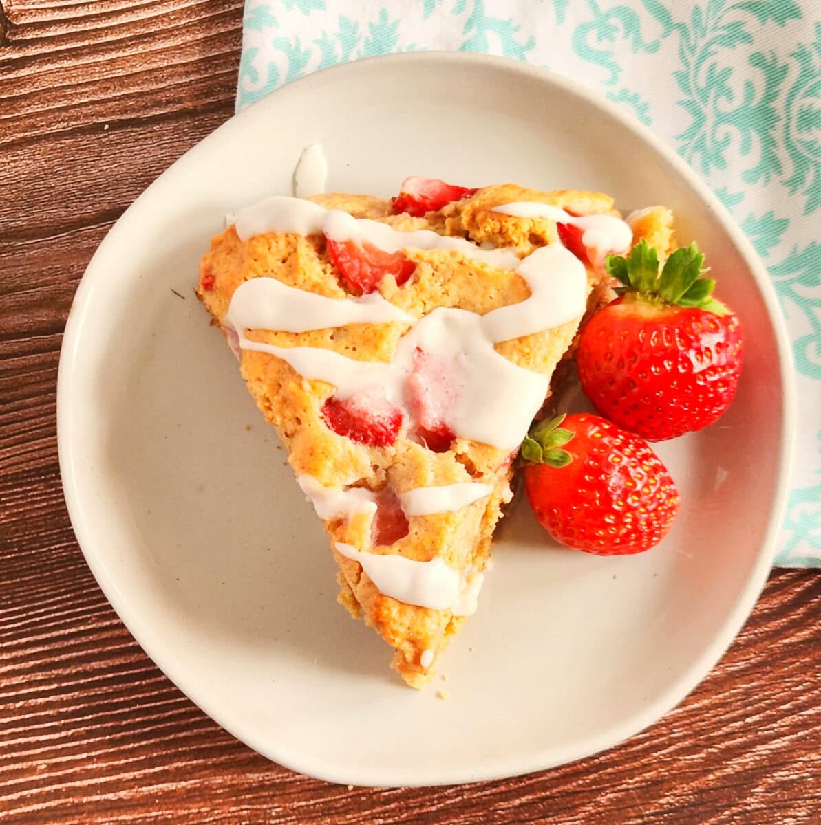 A slice of strawberry scone drizzled with white icing is on a white plate. Two fresh strawberries are beside it. The plate is on a wooden surface with a patterned cloth nearby.