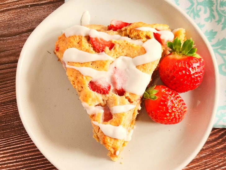 A slice of strawberry scone drizzled with white icing is on a white plate. Two fresh strawberries are beside it. The plate is on a wooden surface with a patterned cloth nearby.