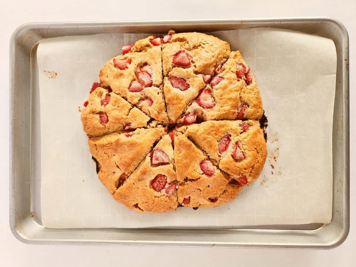 Round baked scone with strawberry pieces, cut into triangular slices, on parchment paper in a baking tray.