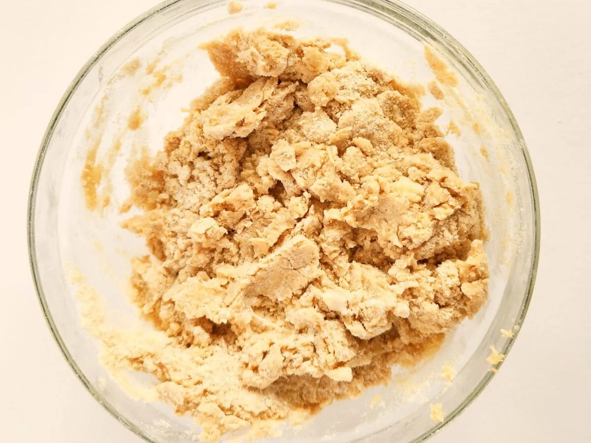 A glass bowl filled with a crumbly, light brown dough mixture on a white background.