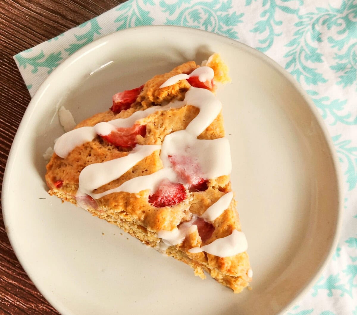 A slice of strawberry scone with icing on a white plate placed on a wooden table with a patterned napkin.