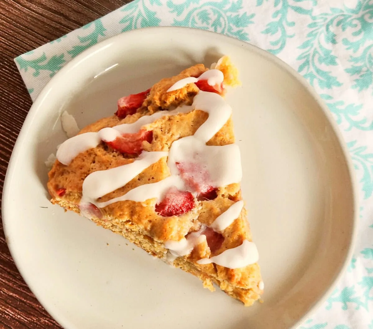 A slice of strawberry scone with icing on a white plate placed on a wooden table with a patterned napkin.