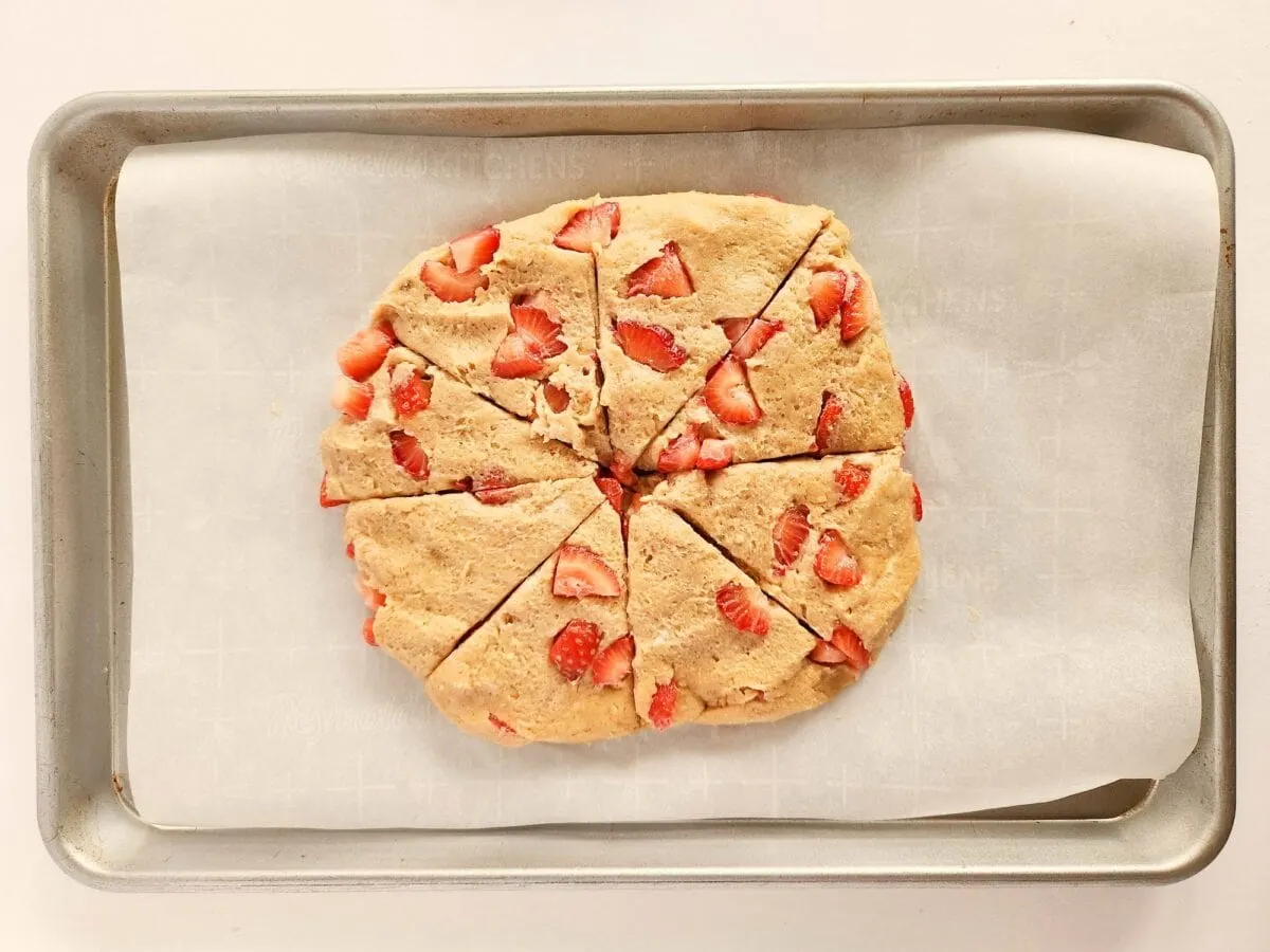 Unbaked scone dough with visible strawberry pieces, cut into wedges, placed on parchment paper on a baking sheet.