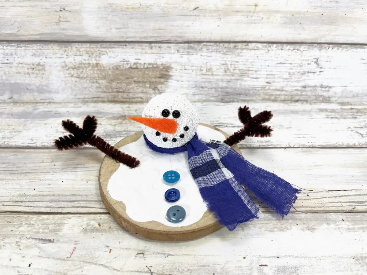 Crafted snowman decoration with a pipe cleaner nose, button eyes, blue checkered scarf, and brown twig arms on a round base, set against a white wooden background.