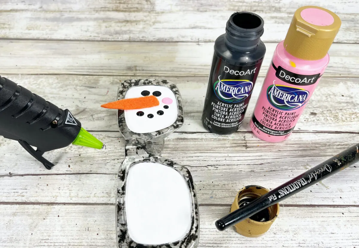 Paint bottles, a hot glue gun, and a painted eyeglass case resembling a snowman with a carrot nose on a wooden surface.