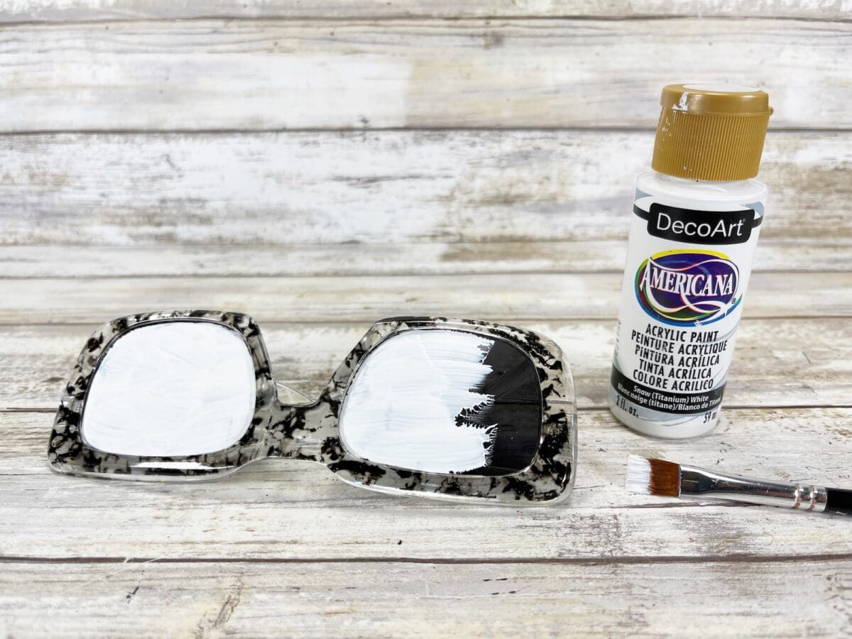 A pair of eyeglass frames partially painted white are on a wooden surface next to a bottle of white acrylic paint and a paintbrush.