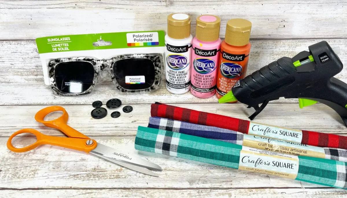 Craft supplies including paint, sunglasses, black buttons, scissors, plaid fabric, and a hot glue gun are arranged on a wooden surface.