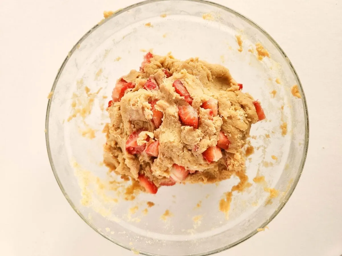 A glass bowl containing cookie dough mixed with chopped strawberries, set against a white background.