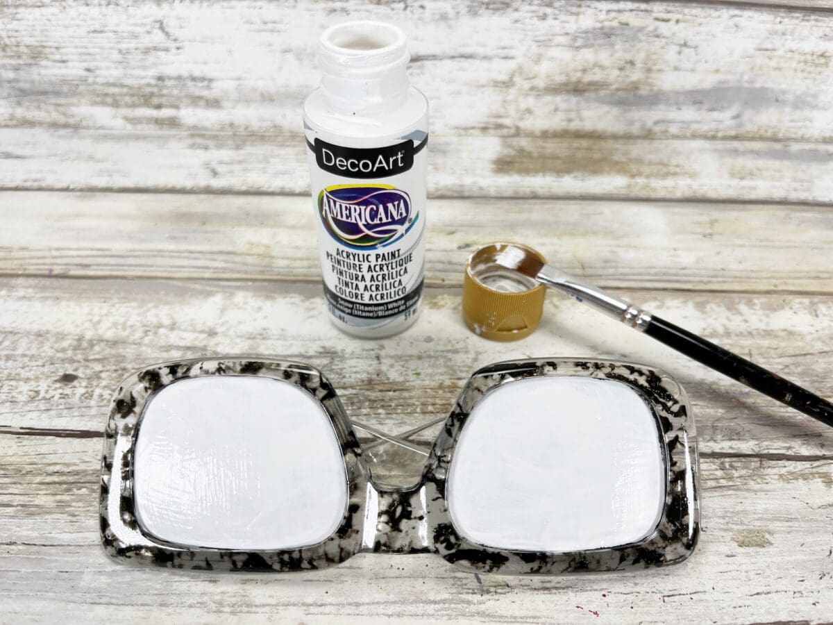 A pair of eyeglasses with white paint on the lenses sits next to an open bottle of DecoArt Americana acrylic paint and a paintbrush on a wooden surface.