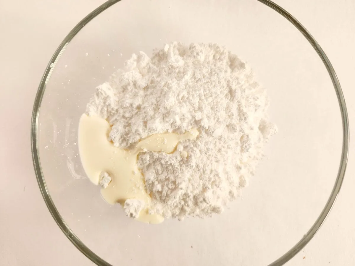 A mixing bowl containing flour and a small amount of liquid on a white surface.