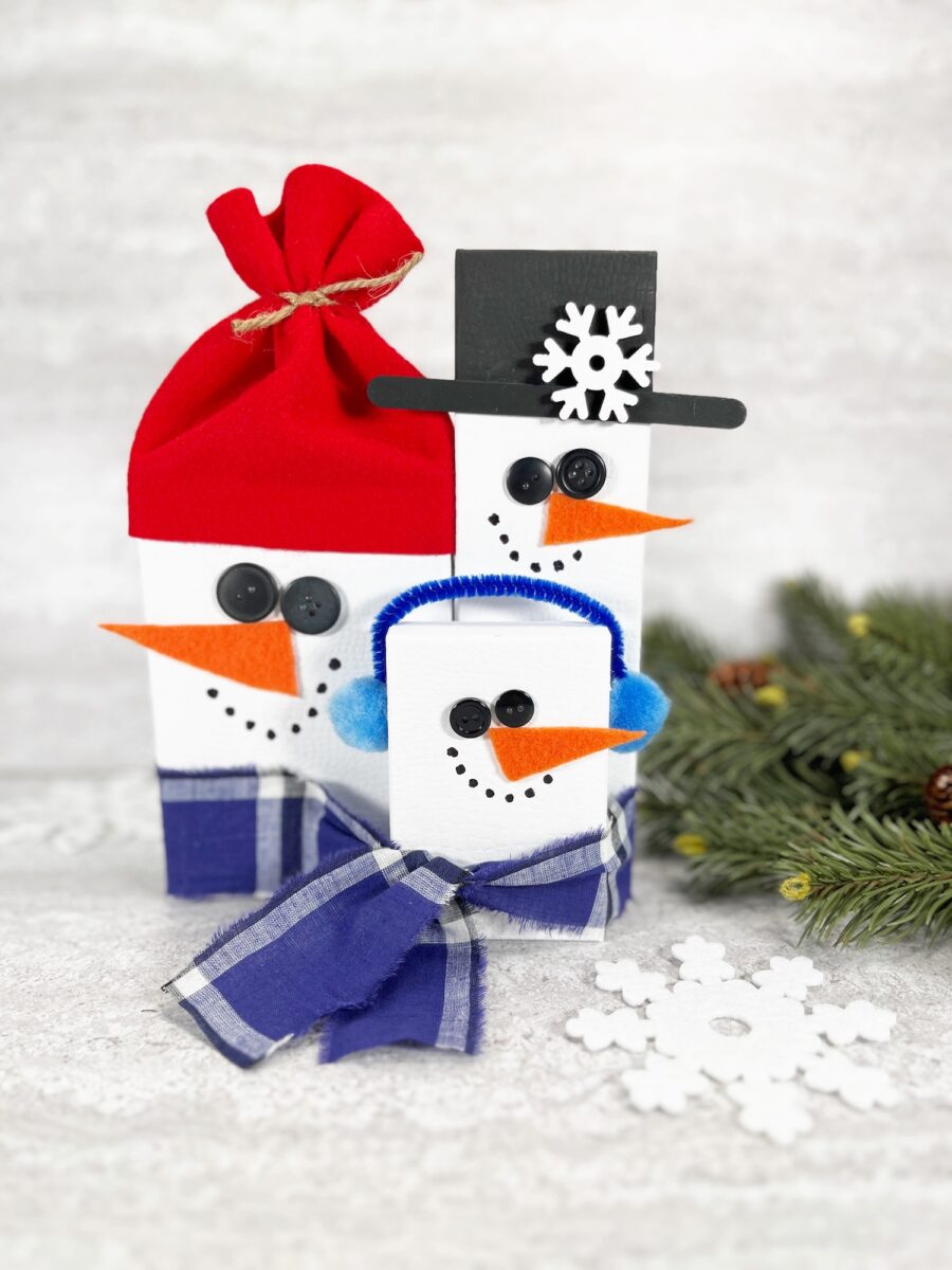 Three snowman-shaped gift boxes with button eyes, carrot noses, and scarves; one wears a red hat, another a black top hat, and the third has blue earmuffs. A pine branch and snowflake nearby.