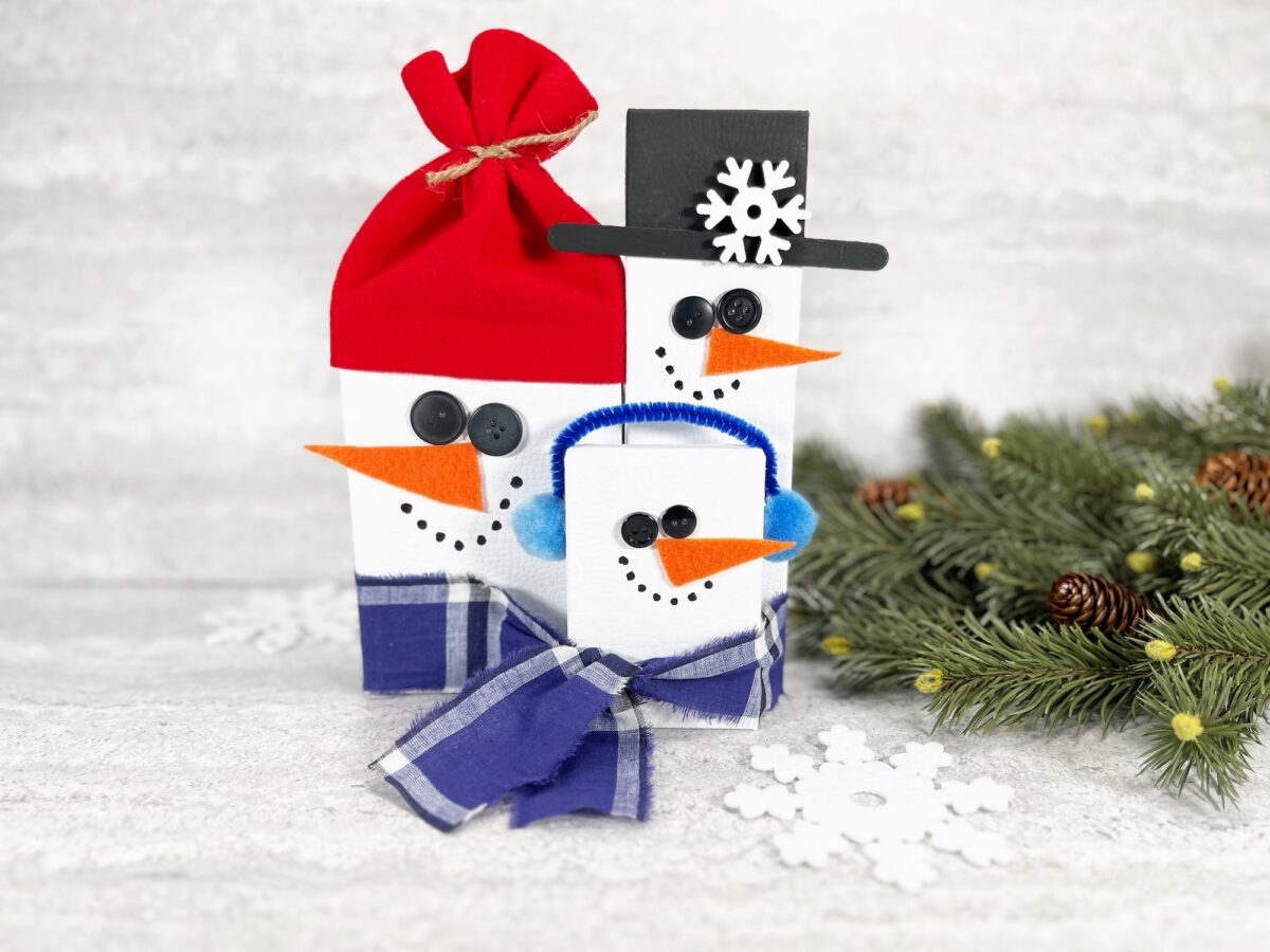 Three snowmen figurines with button eyes and carrot noses, wearing a red hat, black hat, and earmuffs. A pine branch and snowflake decoration are beside them on a white background.