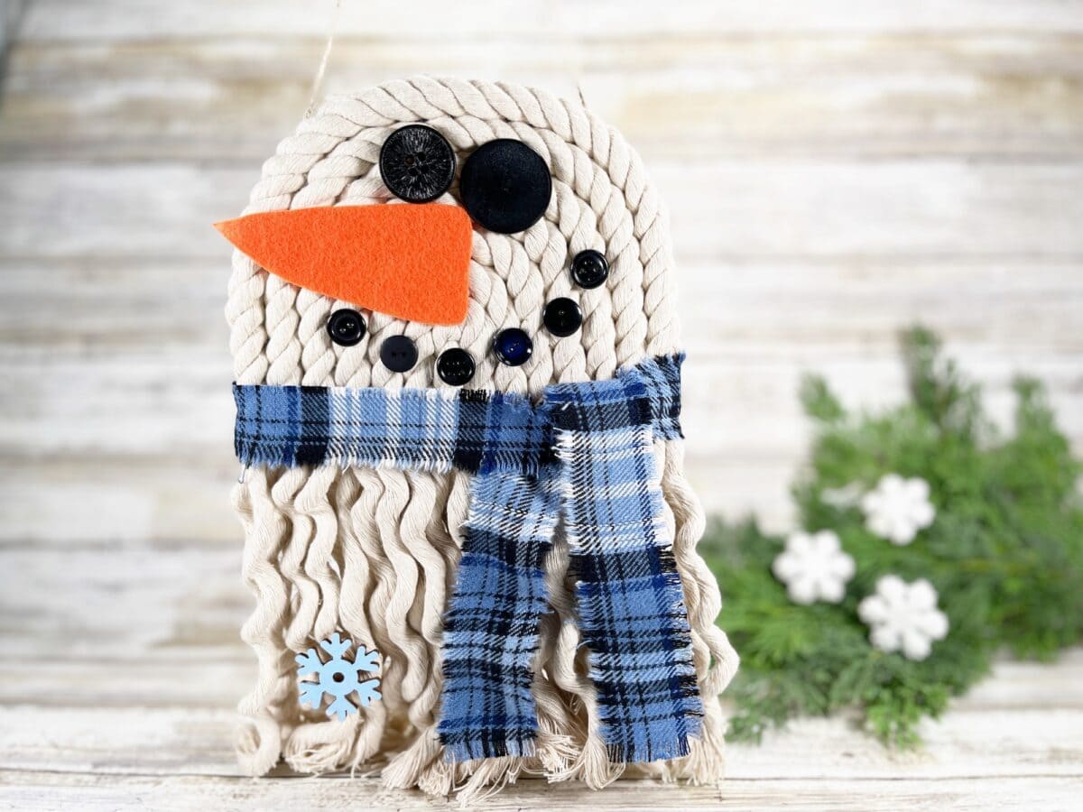 A handmade snowman decoration with a rope face, orange felt nose, button eyes and mouth, blue plaid scarf, and snowflake embellishment, set against a wooden background with greenery.