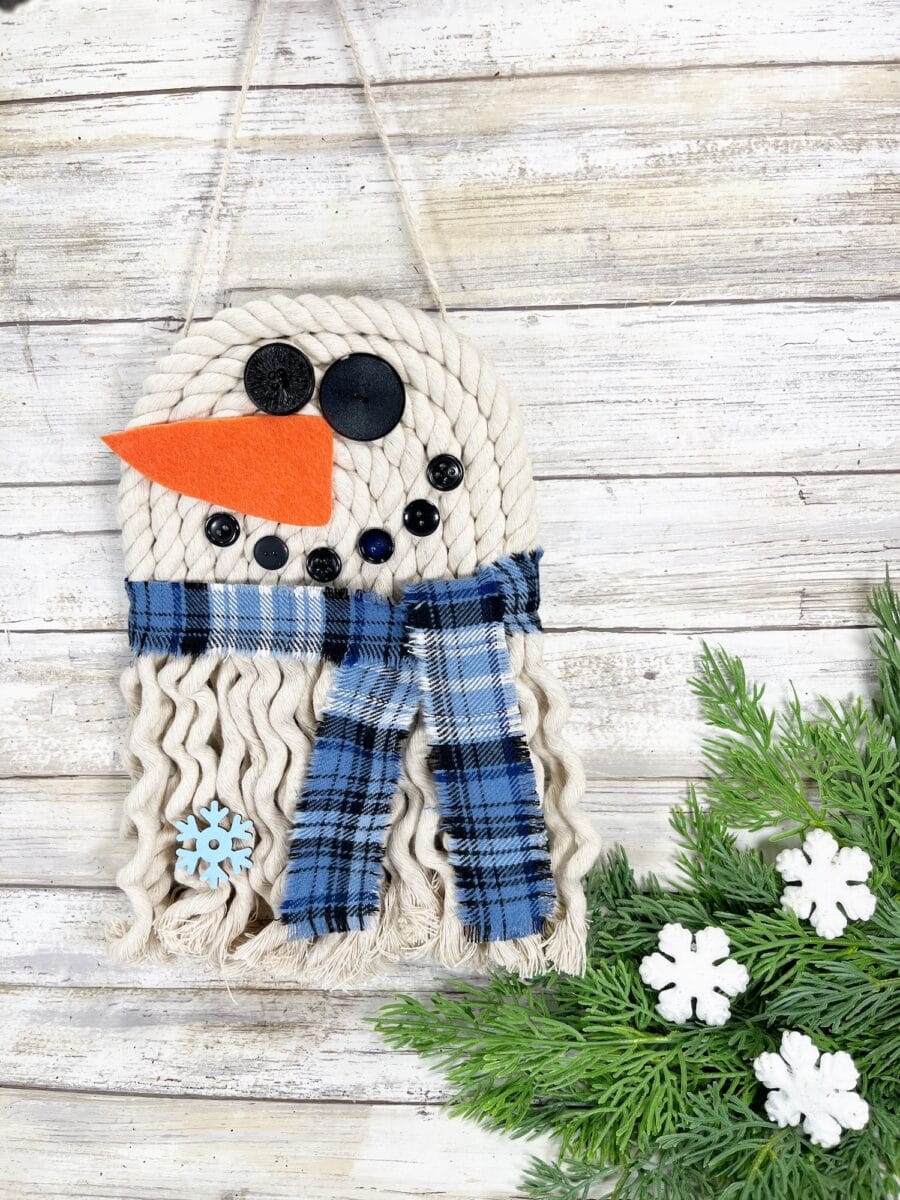 Macrame snowman wall hanging with black button eyes, orange felt nose, and a blue plaid scarf. White wood background with green pine and snowflake decorations on the bottom right.