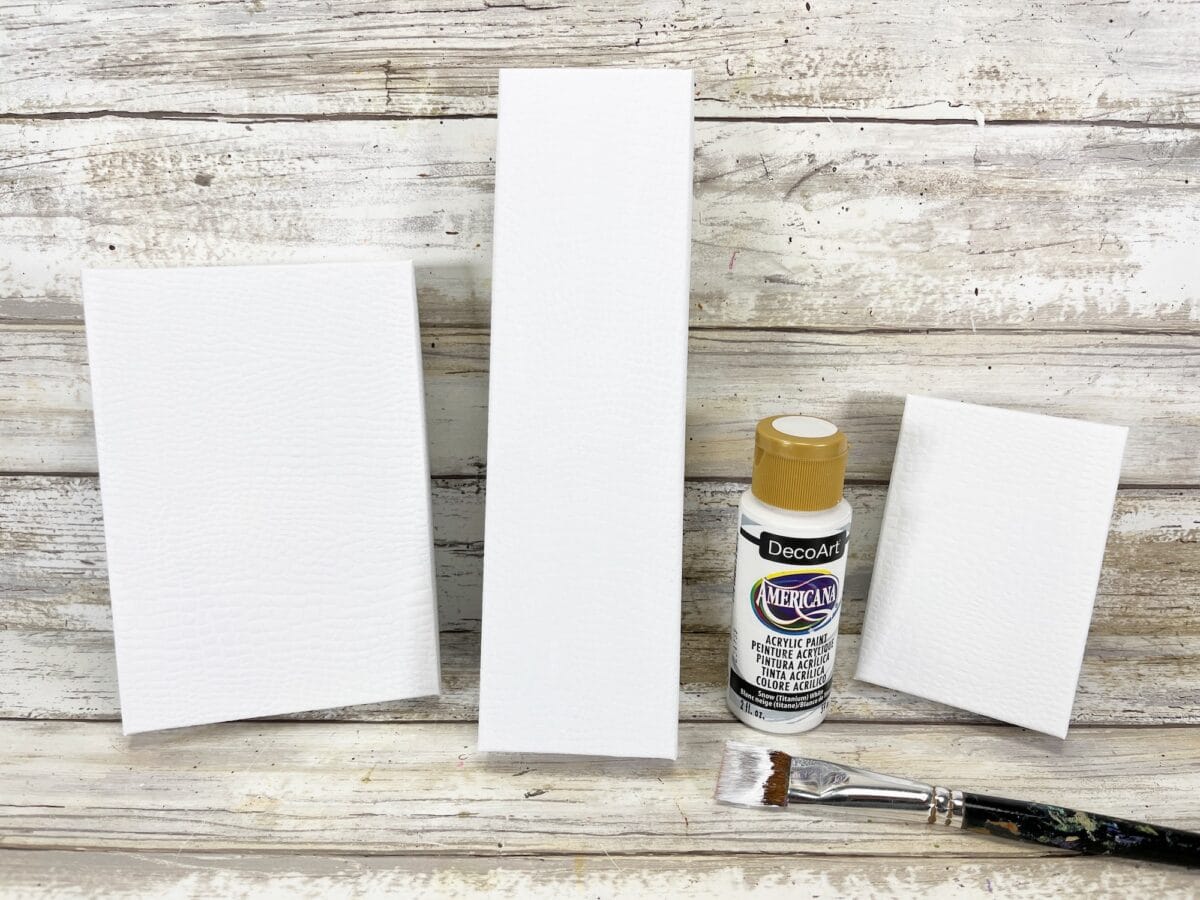 Three blank white rectangular canvases of varying sizes are set on a wooden surface alongside a bottle of tan acrylic paint and a paintbrush.