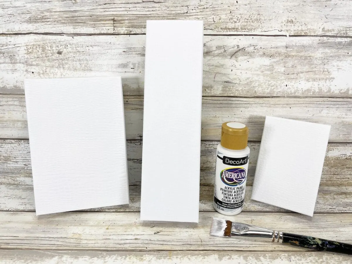 Three blank white rectangular canvases of varying sizes are set on a wooden surface alongside a bottle of tan acrylic paint and a paintbrush.