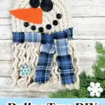 DIY rope snowman with button eyes and carrot nose, wearing a blue plaid scarf. Snowflake decoration and evergreen branch at the bottom.